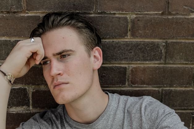 Young adult sits in front of brick wall, grey t-shirt, right hand to forehead, looks pensive