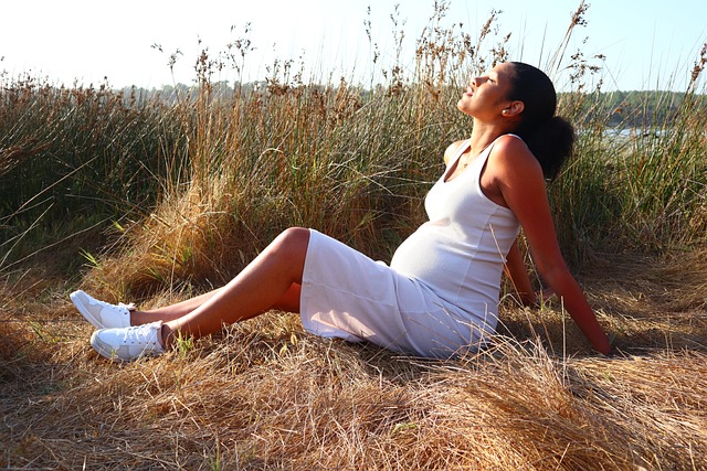 Pregnant woman in white dress sitting on fall grass, looking towards the sun