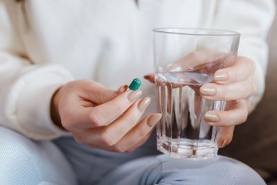 Person holding a green pill and glass of water, only abdomen and part of legs of person are visible