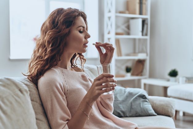 Young woman with long hair sits on couch and has a pill in left hand and a glass of water in right hand