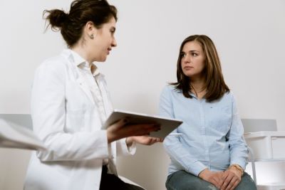 Woman in white smock with clipboard talking to woman in blue shirt