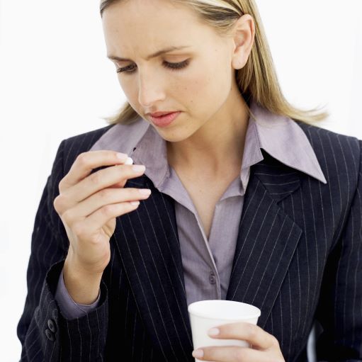 Woman with blond hair, lilac blouse and black striped jacket looking down at a pill in her hand, something to drink in the other hand