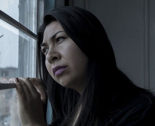 Woman with dark hair looks out of the window, leans head towards window and looks sad