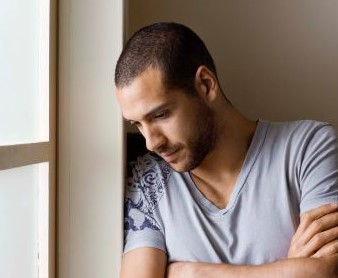 Man with short hair and grey t-shirt standing at window, leaning and looking down