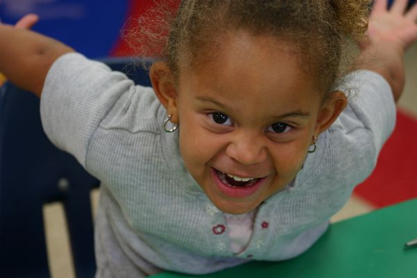 Little girl with grey shirt stretches arm out to back of chair, sits at green table and smiles into camera